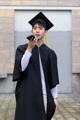Asian teenage male standing in campus courtyard holding smartphone to mouth wearing cap and gown © wavebreak3