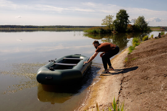 Handsome bearded man pushing green inflatable boat into calm lake water from sandy shore, male fisherman launch process of PVC watercraft, active summer weekend, outdoor recreation concept, copy space