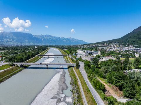 Aerial view of the Rhine River flowing through the valley with bridges and the town of Vaduz surrounded by green trees and mountains under a blue sky in Vaduz, Vaduz, Liechtenstein.
