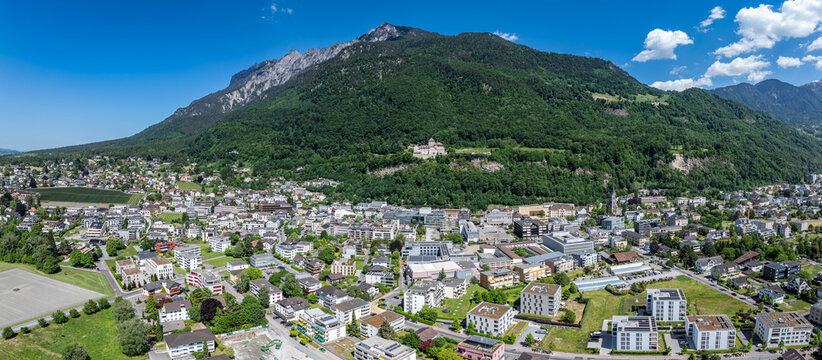Aerial view of Vaduz Castle on a hillside overlooking the city, surrounded by lush green mountains and urban architecture under a clear blue sky in Vaduz, Vaduz, Liechtenstein.