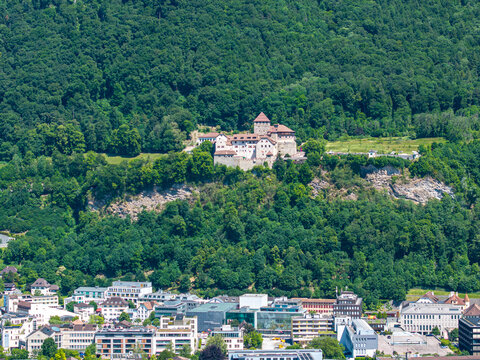 Aerial view of Vaduz Castle perched on a rocky cliff surrounded by lush green forest overlooking the urban buildings of the city below Vaduz, Vaduz, Liechtenstein.