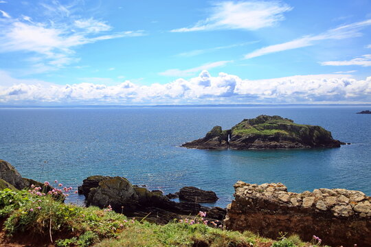 Blick von der K&uuml;ste auf dieArch  &Icirc;le de l'Aber im strahlenden Sonnenschein unter blauem Himmel mit wei&szlig;en Wolken. Im Vordergrund sind Steine und bl&uuml;hende Strand-Grasnelken zu erkennen.