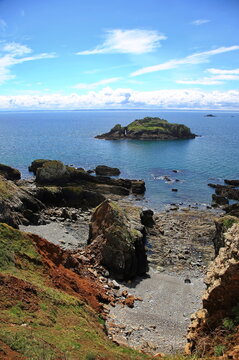 Blick von der K&uuml;ste auf dieArch  &Icirc;le de l'Aber im strahlenden Sonnenschein unter blauem Himmel mit wei&szlig;en Wolken. Im Vordergrund sind Steine und bl&uuml;hende Strand-Grasnelken zu erkennen.