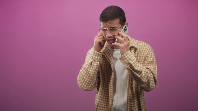 Young hispanic man holds smartphone to ear, pressing ear with fingers and wincing in studio; frustration technical trouble.