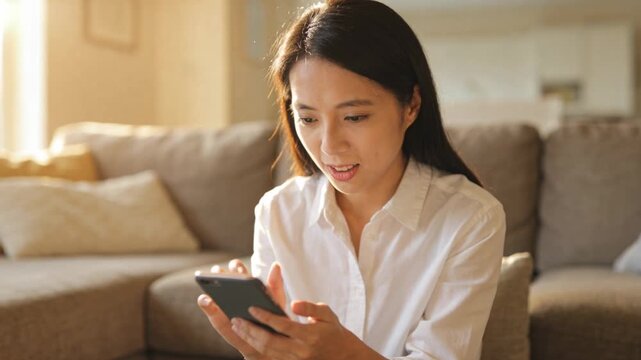 Alert arriving, Asian woman in white shirt tapping smartphone on L-shaped couch, reading message