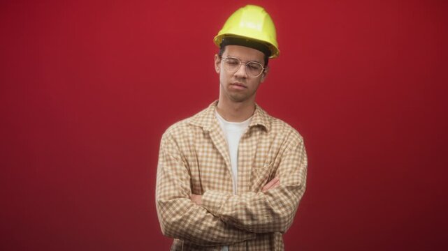 Young hispanic man wearing yellow hardhat and glasses, arms crossed with slight frown against red studio wall; serious focus.