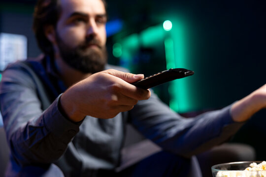 Tired man zapping through channels, watching news broadcast while resting in living room, enjoying leisure. Person using remote control to select TV station, close up shot