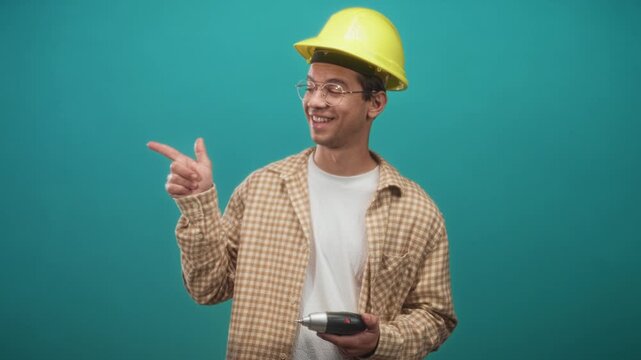 Man builder in yellow hardhat holding drill and points finger to right in studio; confidence skilled.