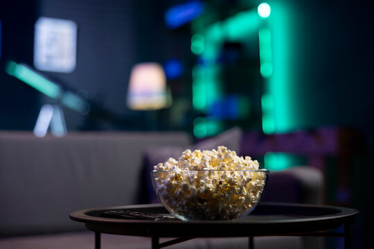 Focus on bowl of popcorn on coffee table in front of cozy couch in empty neon lit modern apartment, blurry background. Close up shot of snack prepared for movie night in home theatre