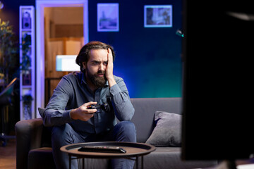 Man looking in disbelief at TV display while playing videogames, receiving game over screen on gaming console. Gamer upset after being defeated by rival players in online esports competition © DC Studio