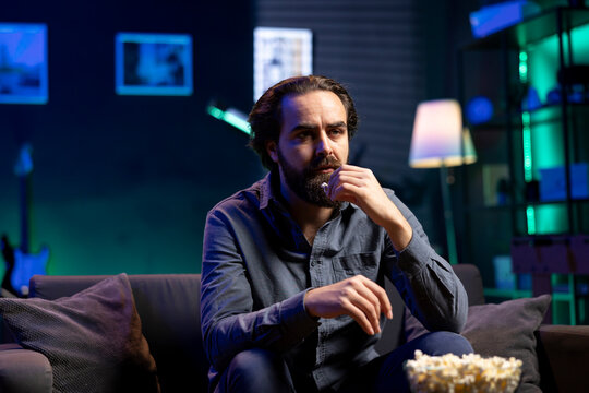 Puzzled man eating popcorn while watching confusing movie on streaming service, enjoying time off from work. Person consuming snacks, looking at films broadcasted on TV with convoluted plots