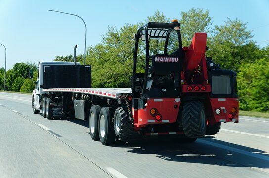 Kent County, Delaware, U.S.A - May 21, 2026 - Flatbed truck transporting a mounted red Manitou forklift along highway under clear sky
