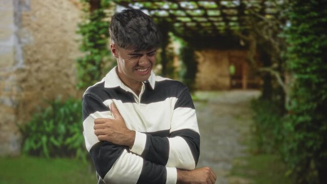 Young hispanic man clutching left shoulder and wincing while gripping his elbow in a vine covered stone building pergola walkway; pain.