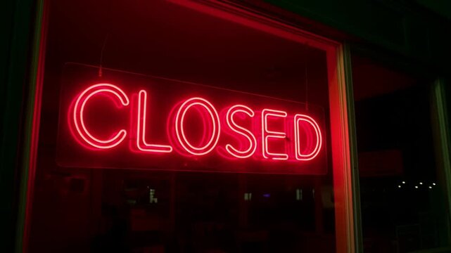 Slow dolly in on neon closed sign glowing and reflecting in storefront window at night with pulsing red light and dark interior