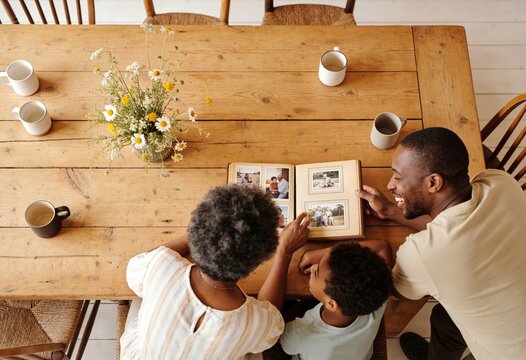 Happy multi-generational Black family looking at a vintage photo album together at a rustic wooden table, grandmother father and son reminiscing about memories at home.