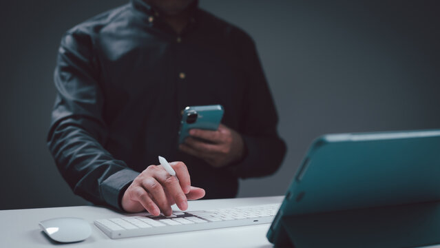 Professional man wearing dark shirt multitasking desk holding smartphone simultaneously typing keyboard next tablet computer stylus mouse dim office studio setting gray background indoors night work.