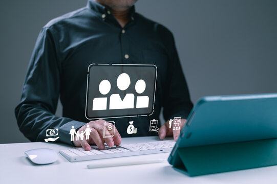 Senior businessman wearing black shirt types computer keyboard office desk managing employee benefits human resources recruitment insurance policy digital icons overlaying workspace environment.