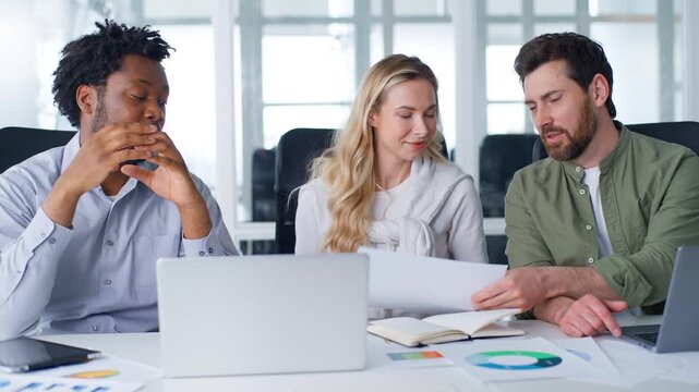 Three diversity business managers in company coworking discuss papers analyzing startup statistic Caucasian businesswoman and businessmen talking looking in document talk work planning office project