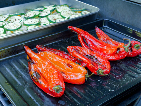 Fresh red pointed peppers and seasoned zucchini slices cooking on a barbecue grill. The scene captures healthy, vegan outdoor cooking during a bright summer day.