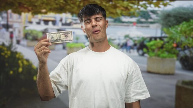 Man holding a fifty dollar bill up near his face, smiling with eyes closed on a bustling street lined with planters and waterfront; playful joy.