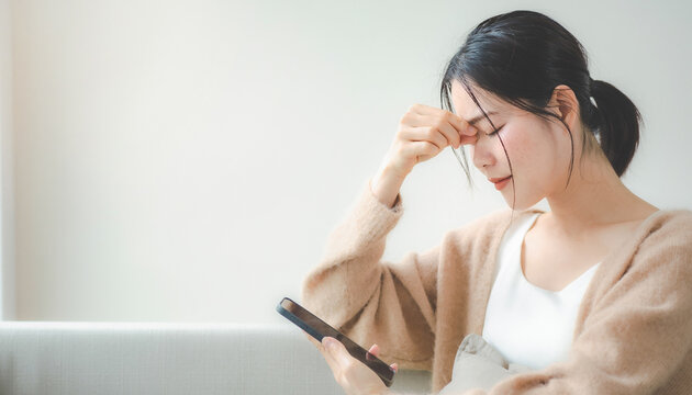Young Asian woman touching nose bridge looking down at smartphone holding head in pain スマホを見ながら目頭を押さえる頭痛や疲れ目に悩む若いアジア人女性