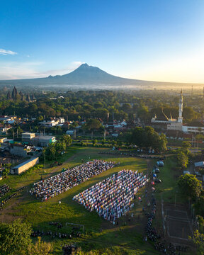 Aerial view of a large crowd gathered for morning prayer in an open field with Mount Merapi and Prambanan Temple under a clear sky in Indonesia.