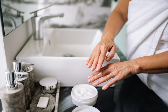 Close-up of a senior woman's hands in a bathroom, near a sink. She is carefully applying hand cream to her dry, wrinkled skin, seeking moisture and gentle aging care as part of her routine