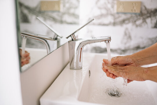 Mature woman washes her hands under running water at a modern bathroom sink. The chrome faucet dispenses a steady stream into her cupped hands, promoting clean health and daily personal hygiene