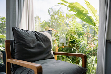 A comfortable grey armchair with a wooden frame sits by a large window. Outside, bright sunlight illuminates dense tropical foliage, creating an ideal spot for relaxation and rest on a calm day