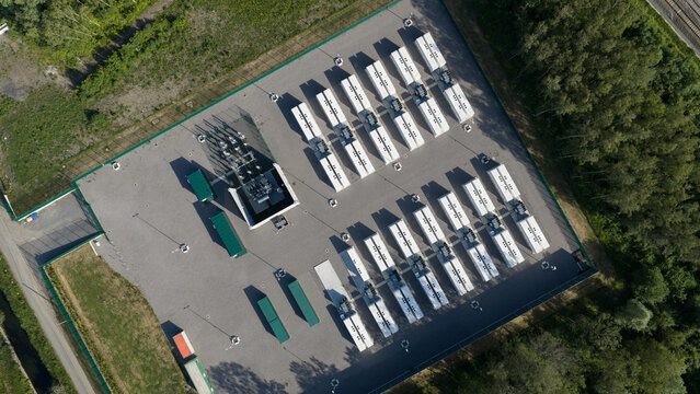 Aerial view of rows of white battery storage units and electrical infrastructure on a gravel lot surrounded by green trees in Le Roeulx, Wallonia, Belgium.