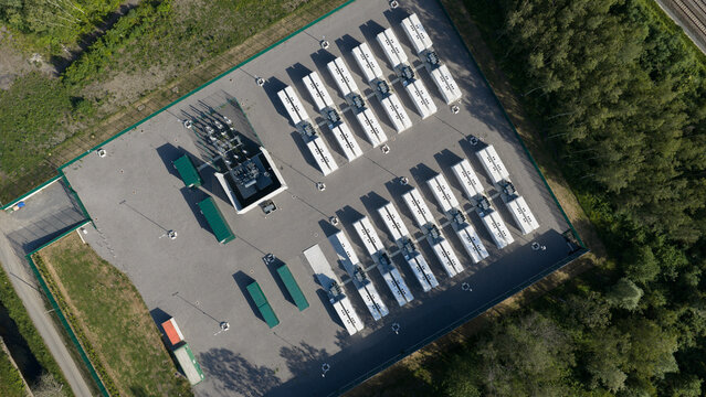 Aerial view of a battery energy storage system with rows of white container units and electrical infrastructure surrounded by lush green forest in Le Roeulx, Wallonia, Belgium.