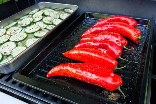 Fresh red pointed peppers and seasoned zucchini slices are cooking on a barbecue grill. A close-up of a gas grill loaded with fresh vegetables.