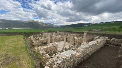Fototapeta premium Ancient ruins of Satala Ancient City in Kelkit, Gümüşhane, Türkiye. Historic Roman archaeological site featuring stone foundations and columns set against a dramatic cloudy sky and lush green landscap