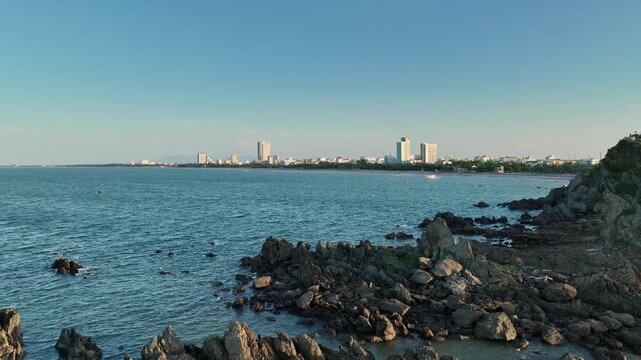 Rocky coastal foreground beside Lan Chau Island with calm sea waters and distant buildings under clear morning light.