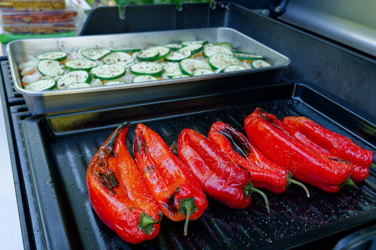 Fresh red pointed peppers and seasoned zucchini slices are cooking on a barbecue grill. A close-up of a gas grill loaded with fresh vegetables.