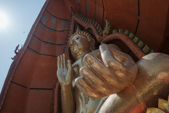 A low-angle, close-up shot focusing on the hand and face of a massive golden Buddha statue at Wat Tham Suea, the famous Tiger Cave Temple in Kanchanaburi, Thailand, set against a clear blue sky.
