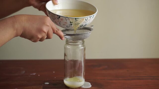 Close up hand pouring melted beef fat (tallow) from bowl into glass jar on wooden table, homemade tallow storage process
