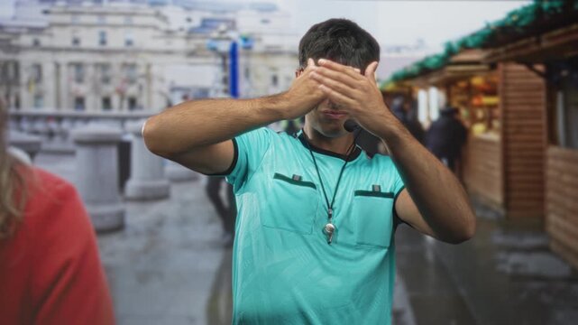 Young referee man covering face with hand and holding whistle on lanyard in street; privacy withdrawal.