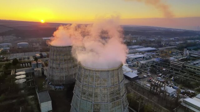 Aerial view of cooling towers and power plant smoke stack emitting thick vapor clouds during sunset.