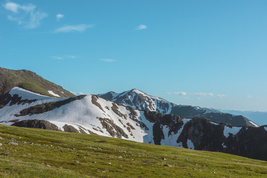 Scenic alpine view from grassy hilly meadow above abyss to big snowy rocky mountain ridge under clouds in blue sky in sunny day. Colorful upland landscape with large rocks with snows in bright sun.