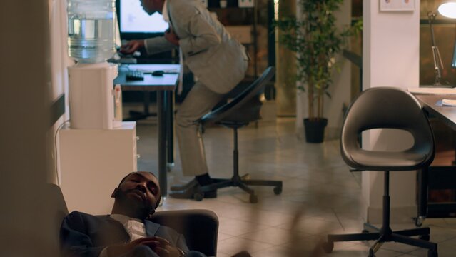 Team leader finishing nightshift, ready to go home, catching tired assistant taking nap on office couch. Manager annoyed by employee slacking off by sleeping in workspace