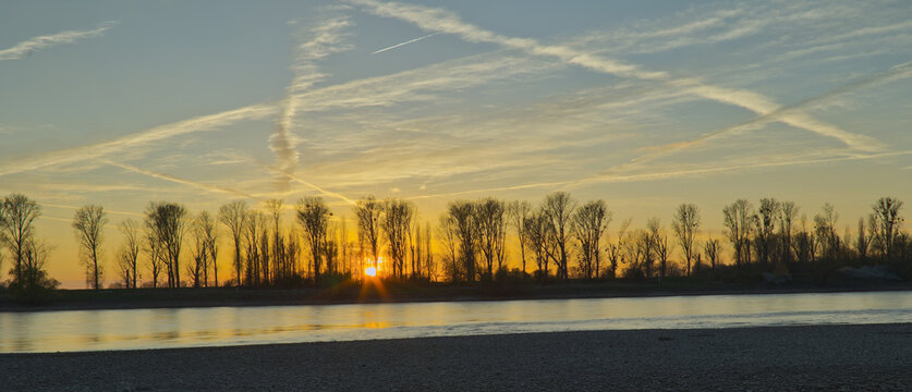 Ruhige Flusslandschaft am Rhein im Gegenlicht der untergehenden Sonne und kahlen Pappeln im Hintergrund