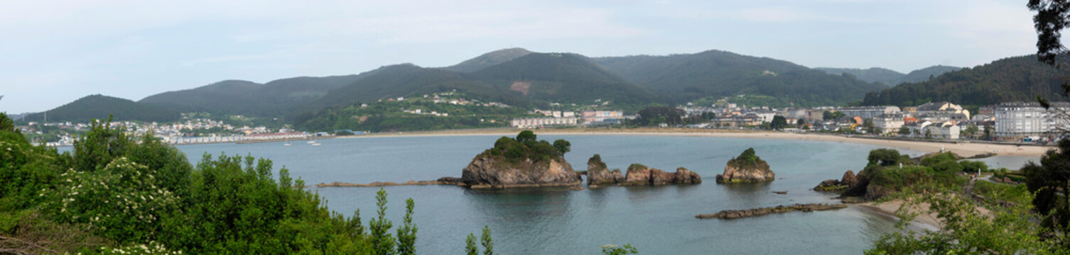 Os Castelos rock formations on Covas Beach in Viveiro, Galicia, showcasing mossy coastal stones, seagulls on the sand, and a hillside town in the background.