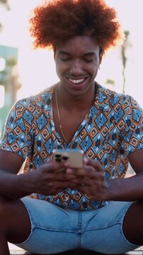 Happy and carefree African American male browsing website on mobile phone while
sitting outdoors in summer city. Smiling smartphone user downloads application in
store. Hipster guy having video call