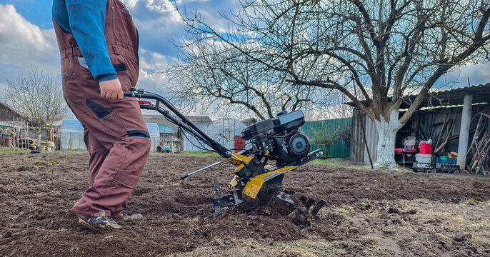 Man operating a walk-behind tractor, tilling and cultivating spring garden soil in a backyard plot on a cloudy day, preparing ground for planting and homestead work