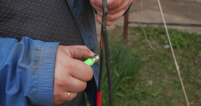Close-up of an electrician's hands repairing a broken electrical wire outdoors by heating and shrinking insulation tubing with a lighter to create a secure and waterproof connection