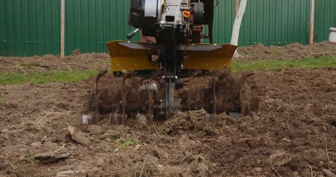 Male farmer using a garden rototiller to cultivate the soil before planting. Agricultural machinery preparing the land for seeding in a field, with rotating blades digging into the ground