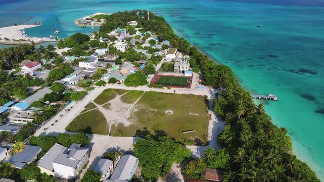 Aerial view of the tropical island of Fulhadhoo featuring a village, lush palm trees, and a large open field by turquoise ocean waters in Fulhadhoo, Baa Atoll, Maldives.