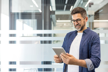 Fototapeta premium Smiling man wearing glasses and a denim shirt standing in a modern office, engaged with a digital tablet, representing mobile technology, communication, and business connectivity
