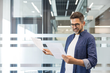 Fototapeta premium Confident man reviewing corporate paperwork in a contemporary office setting, focusing on data analysis, planning, and achieving business goals with a positive expression